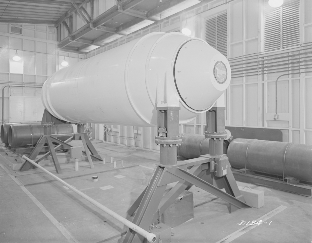 Ein Schwarz-Weiß-Foto einer großen zylindrischen Wasserstoffbombe auf einem Ständer in einer Fabrik mit Rohren, Lampen und einer Wand im Hintergrund.