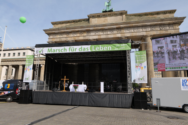 Bühne vor dem Brandenburger Tor mit einem Tisch, Bannern, Lautsprechern, Fahrzeugen, Gebäuden, einer Statue, einer Flagge und einer bewölkten Himmel.