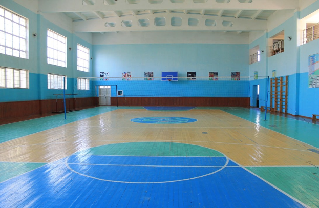 Große Turnhalle mit blau-weißem Boden, einem Basketballfeld, einem Netz mit Stangen, einer Tür, Fenstern, Wandplakaten, einem Geländer und einem Dach.
