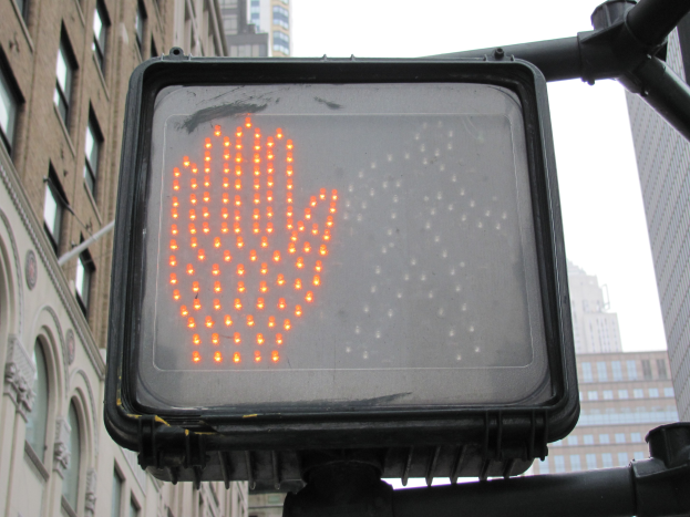 Rote Hand am Verkehrssignal in einer städtischen Umgebung mit hohen Gebäuden auf beiden Seiten und einem klaren Himmel darüber.
