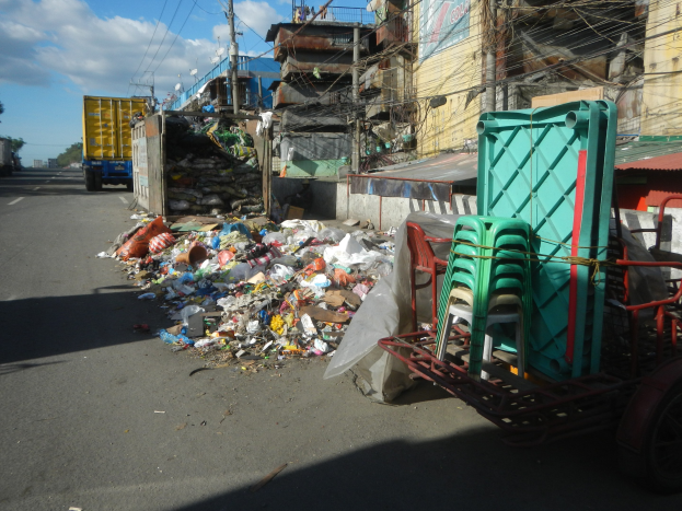 Ein Lastwagen neben einem Müllhaufen auf einer Straße, mit einem Einkaufswagen mit Plastikstühlen rechts daneben und Gebäuden, Strommasten, Bäumen und einem bewölkten Himmel im Hintergrund.