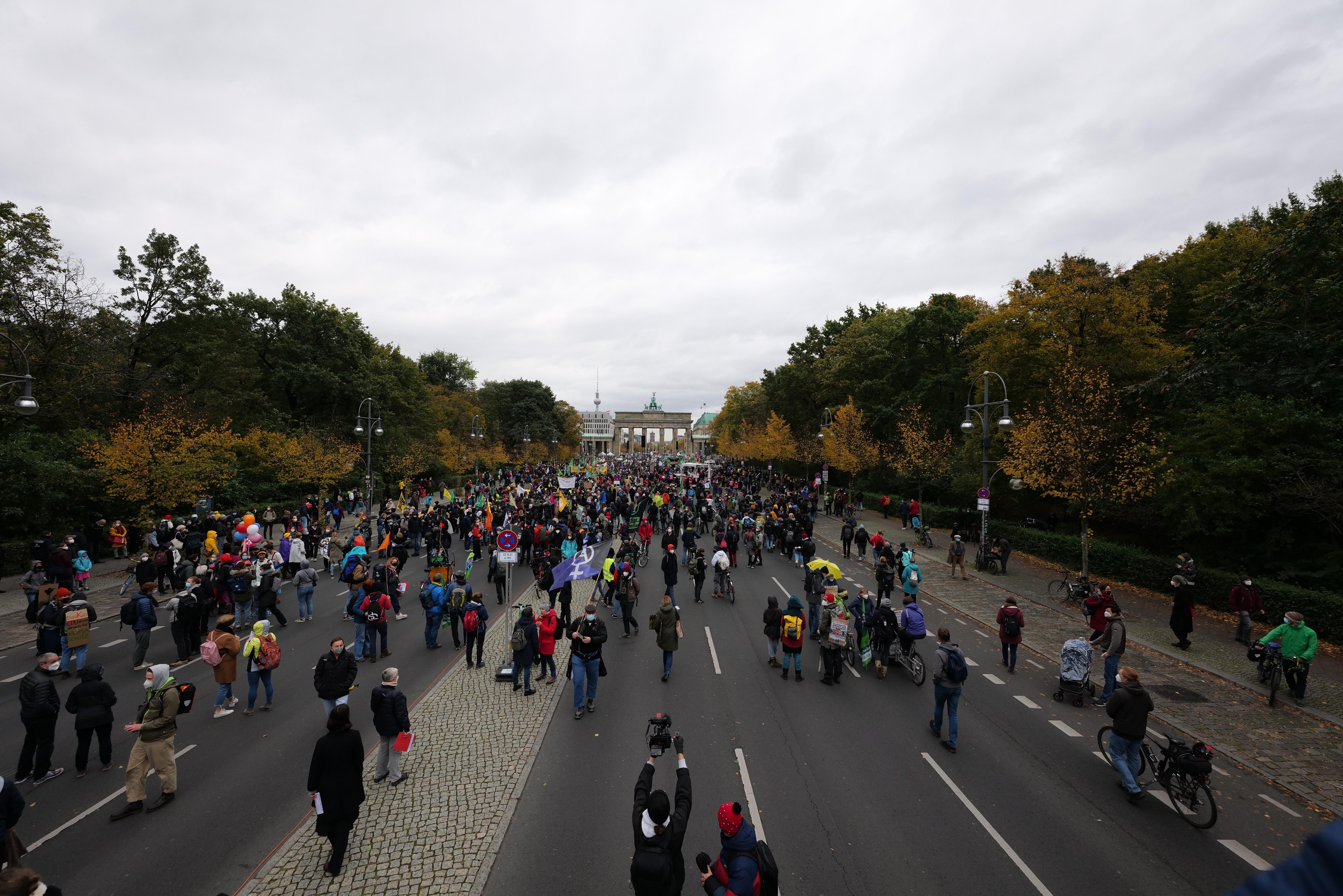 Eine große Gruppe von Menschen marschiert eine von Bäumen gesäumte Straße mit Laternenpfählen entlang, mit Kameras in der Hand, mit einem Gebäude und einem klaren Himmel im Hintergrund.
