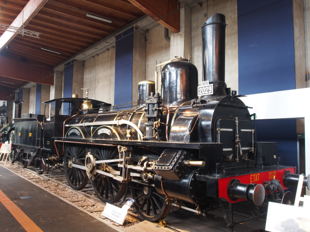 Alte Dampflokomotive im Museum ausgestellt, umgeben von Eisenbahnschienen, Informationsschildern und anderen Ausstellungsstücken, mit beleuchteter Decke im Hintergrund.