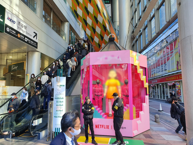 Eine Gruppe von Menschen steht vor einem gläsernen Gebäude mit Texttafeln, einige tragen Masken, während eine Rolltreppe mit Fahrgästen zu sehen ist; ein pinker Kasten in der Mitte zeigt Schaufensterpuppen und trägt den Text "Netflix's new Netflix booth in Tokyo."