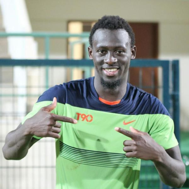 Ein Mann in einem grünen und blauen Hemd, der in die Kamera lächelt und darauf zeigt, mit einem unscharfen Geländer, Wand und Tür im Hintergrund.