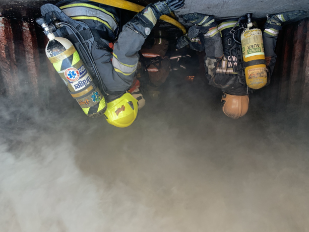 Feuerwehrleute in Schutzausrüstung löschen einen Tunnelbrand mit Rauch, der aus der Tiefe aufsteigt.