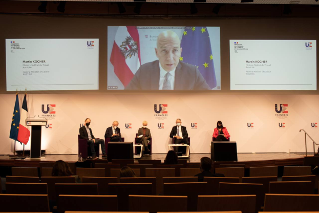 Eine Gruppe von Menschen, die auf einer Bühne in einem Halbkreis um ein zentrales Podium mit Mikrofon sitzen, flankiert von Flaggen, mit einem Bildschirm im Hintergrund, der Text und Bilder anzeigt, während einer Pressekonferenz der Europäischen Union.