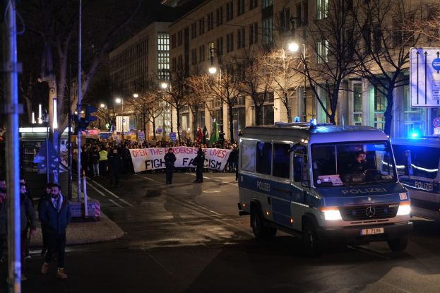 Eine Gruppe von Menschen, die nachts die Straße entlanggehen, mit einem Polizeiwagen auf der Seite und einer im Hintergrund sichtbaren Fahne.
