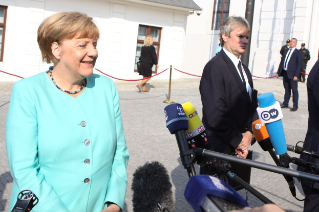 Bundeskanzlerin Angela Merkel spricht mit Reportern vor einem Gebäude mit einem bewölkten Himmel, umgeben von Menschen und Mikrofonstativen mit Ständern, mit einem Seil, das an einem Pfahl befestigt ist.