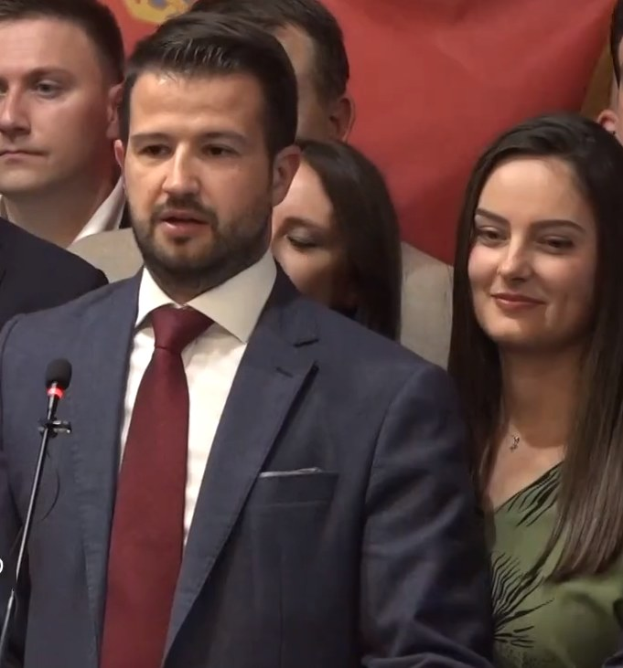 Ein Mann im Anzug spricht in ein Mikrofon an einem Rednerpult, mit einem Banner dahinter und einer Menge davor, wahrscheinlich zu einem Regierungsverbot der sozialen Medien.