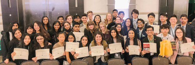 Gruppe von Menschen mit Urkunden in einer Abschlussfeier-Situation, umgeben von Stühlen und einer Wand im Hintergrund.