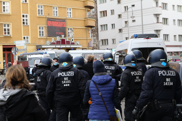 Polizeibeamte in Uniform vor einer Menge mit Helmen und Jacken während einer Demonstration in Berlin, Deutschland, mit Fahrzeugen, Gebäuden, Laternenpfählen und einem Banner im Hintergrund.