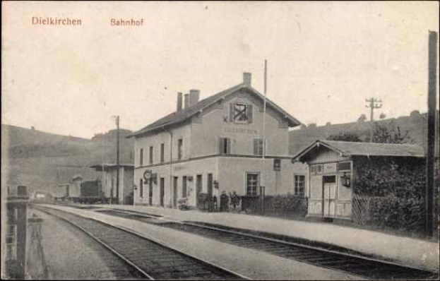 Schwarzes und weißes Foto einer Bahnstation in Dielkirchen, Deutschland, mit einem Zug auf den Gleisen, umgeben von Häusern, Strommasten, Bäumen und Menschen, mit Hügeln und einem klaren Himmel im Hintergrund.