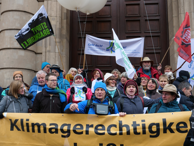 Eine Gruppe von Menschen vor einem Gebäude mit Säulen und einer Tür, die ein Transparent mit Text, Fahnen mit Stangen und Luftballons halten, nehmen an einem Klimastreik in Deutschland teil.