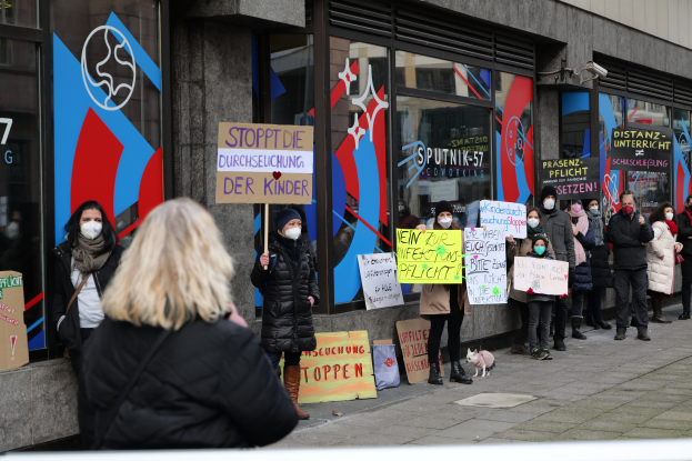 Gruppe von maskierten Menschen mit Schildern und Plakaten vor einem gläserummantelten Gebäude, mit Kameras und einem Hund, protestieren gegen eine deutsche Regierungentscheidung, Masken in Schulen zu verbieten.