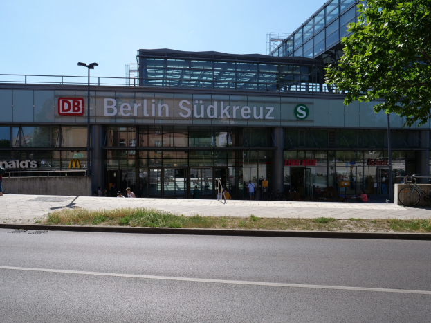 Großes glaswandiges Berliner Südkreuz-Bahnhof mit sichtbarem Text, umgeben von Straßenmasten, Lampen, Fahrzeugen, Fußgängern, Fahrrädern, Bäumen und einem klaren blauen Himmel.