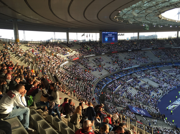 Große Zuschauermenge in einem Stadion bei einem Fötballspiel, mit einer Bühne auf der rechten Seite, Fahnen, Masten, einem Bildschirm und der Allianz Arena in München im Hintergrund.