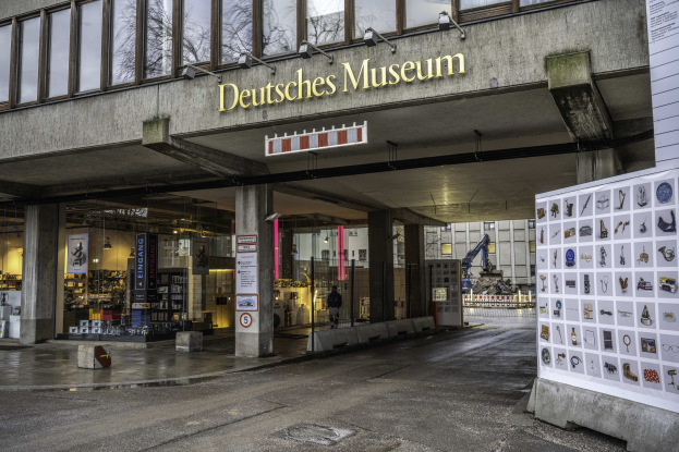 Großes Gebäude mit Glasfenstern, Säulen und einer Namenstafel mit der Aufschrift "Deutsches Museum" in Berlin, Deutschland, mit einer Anzeigetafel mit Bildern und Text auf der rechten Seite, vor dem Hintergrund anderer Gebäude, Bäume und Himmel.