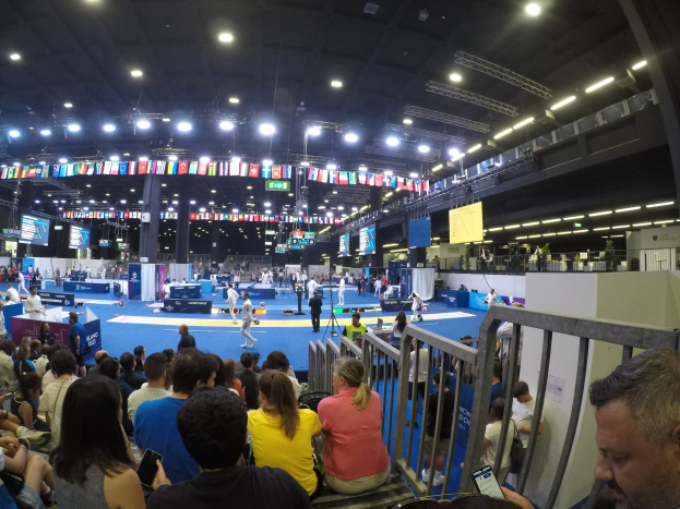 Große Menschenmenge in einer Indoor-Sportarena vor einer Bühne mit stehenden Zuschauern, Bildschirmen, Fahnen und Deckenleuchten im Hintergrund.