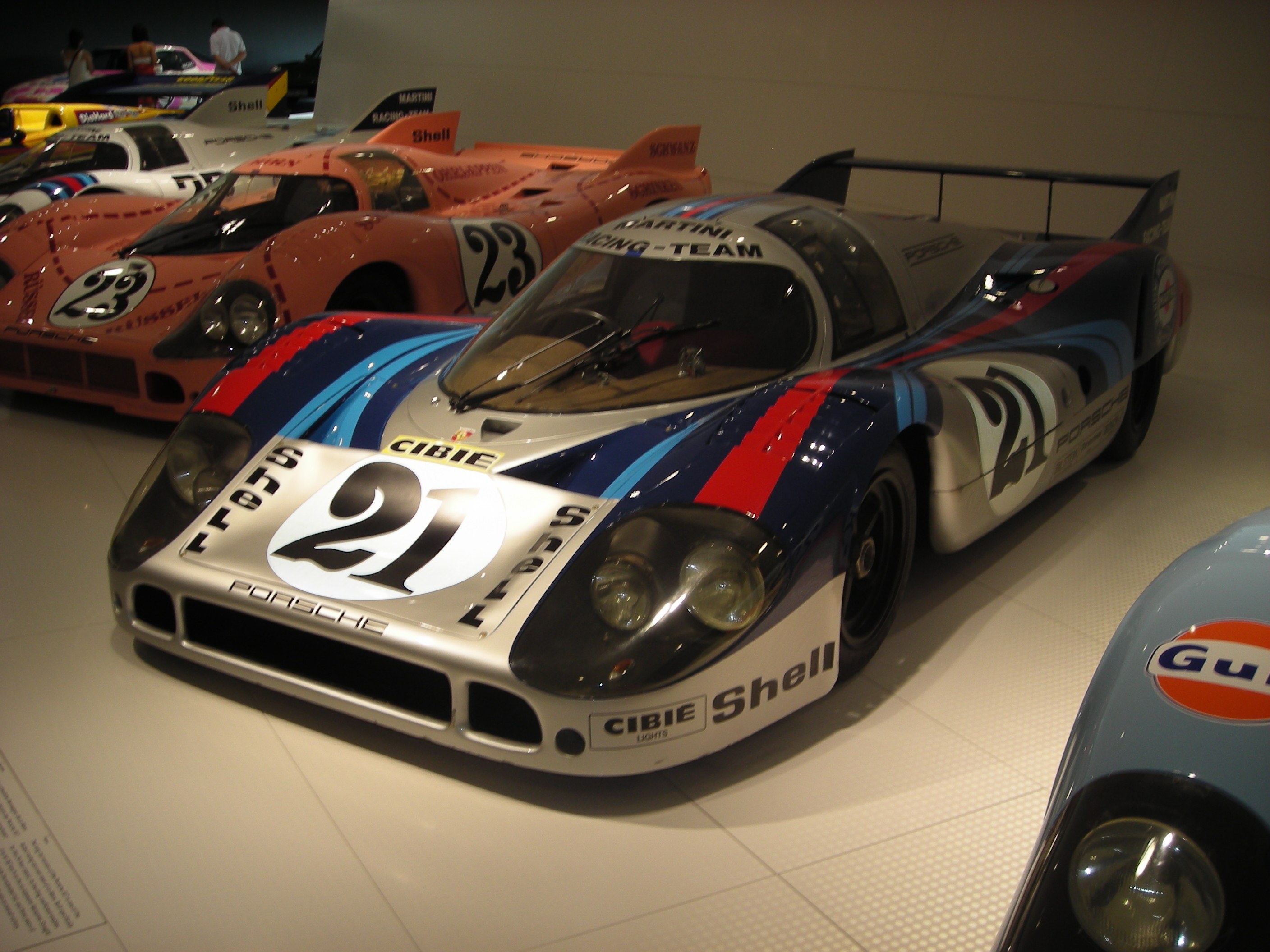 Ein Porsche 917K Le Mans-Rennwagen, der in einem Museum ausgestellt wird, umgeben von anderen Sportwagen auf dem Boden, mit Menschen in der Nähe und einer Wand im Hintergrund, zusammen mit einem Textplakat auf der linken Seite.