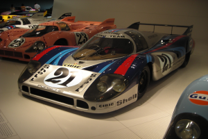 Ein Porsche 917K Le Mans-Rennwagen, der in einem Museum ausgestellt wird, umgeben von anderen Sportwagen auf dem Boden, mit Menschen in der Nähe und einer Wand im Hintergrund, zusammen mit einem Textplakat auf der linken Seite.