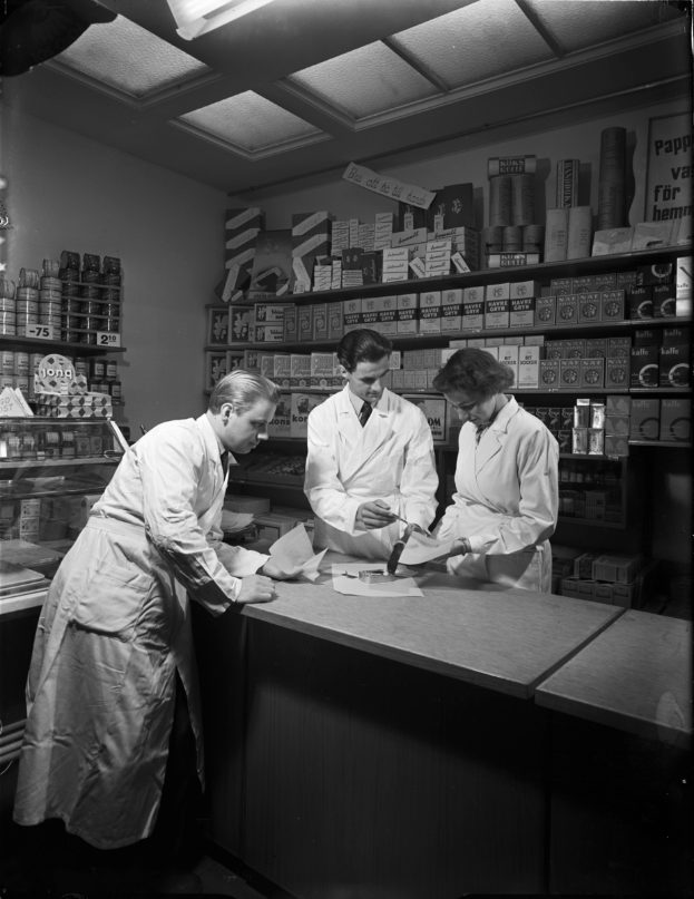 Drei Männer in weißen Kitteln stehen an einem Tresen in einer Apotheke mit Papieren und Gegenständen darauf, mit Regalen im Hintergrund, dargestellt in Schwarz-Weiß.