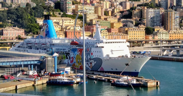 Großes Kreuzfahrtschiff liegt in der Nähe einer Stadt mit einer Brücke auf der linken Seite, Fahrzeugen auf der Straße und Gebäuden und Bäumen im Hintergrund.