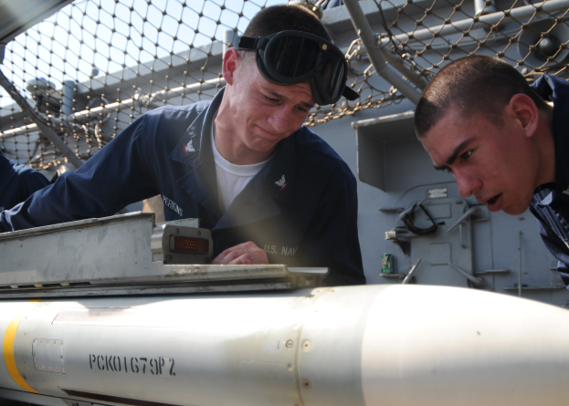 Zwei Männer in Marineuniformen arbeiten an einer Rakete auf einem Schiff, umgeben von einem Metallzaun und einem klaren blauen Himmel.