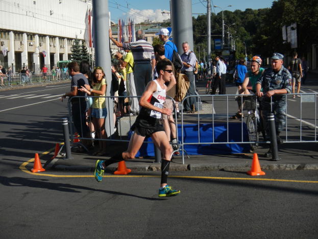 Ein Mann läuft bei einem Marathon auf einer Stadtstraße, umgeben von Zuschauern mit Fahrrädern, mit städtischer Infrastruktur und einem bewölkten Himmel im Hintergrund.