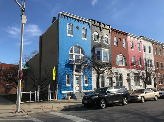 Eine Reihe bunter Häuser an einer Straßenecke in Baltimore, Maryland, mit parkenden Autos, Bäumen, Strommasten, einem Zaun und einem bewölkten Himmel.