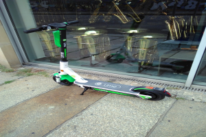 Elektro-Scooter auf einem Gehweg vor einem Laden mit einer Glaswand, die Musikinstrumente und andere Gegenstände zeigt.