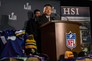 Ein Mann in einem schwarzen Blazer, wei├čeem Hemd und schwarzer Krawatte spricht an einem Pult mit Mikrofon und Logo vor einer Menge. Blaue und gelbe Merchandising-Artikel und ein "Super Bowl LII"-Banner sind im Hintergrund zu sehen.