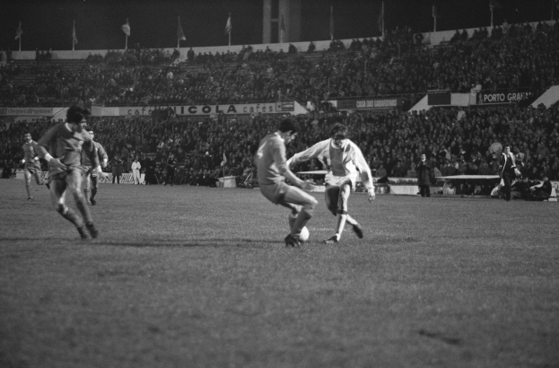 Schwarzes und weißes Foto von Männern, die Fußball in einem Stadion mit Zuschauern auf den Rängen und Fahnen im Hintergrund spielen.