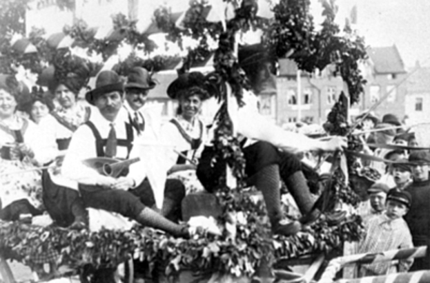 Schwarz-weißes Bild von Menschen in traditioneller bayrischer Tracht auf einem geschmückten Paradewagen, einige spielen Musikinstrumente, mit Gebäuden und einem klaren Himmel im Hintergrund.