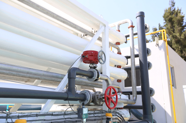 Große Gruppe von Rohren, Ventilen, Metallstangen und anderen Komponenten auf einem Gebäudedach mit Bäumen und einem klaren blauen Himmel im Hintergrund, die die Technologie der Wasseraufbereitungsanlage darstellen.