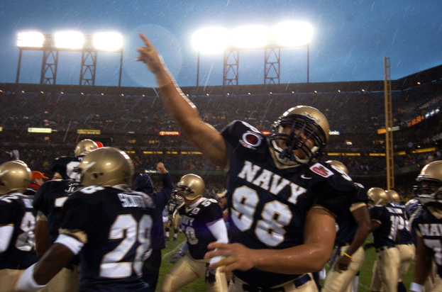 Navy-Football-Spieler feiern Touchdown bei Regen auf einem nassen Feld, tragen Helme und Uniformen, mit Zuschauern und Stadionbeleuchtung im Hintergrund.