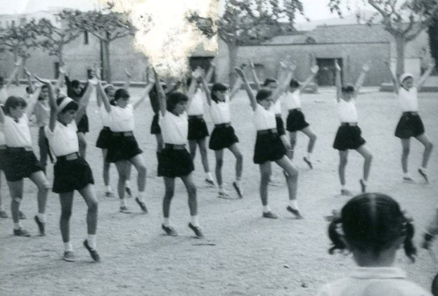 Schwarzes und weißes Foto von jungen Mädchen in Schuluniformen, die auf einer Straße tanzen, die von Gebäuden und Bäumen gesäumt ist, mit einer Person in Schuhen unten sichtbar.