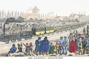 Gruppe von Menschen um einen Zug auf Schienen mit einigen Passagieren im Inneren und anderen draußen, vor Gebäuden und Bäumen mit Text unten lesend "Ludwigs-Eisenbahn, der erste Bahnhof in Deutschland."