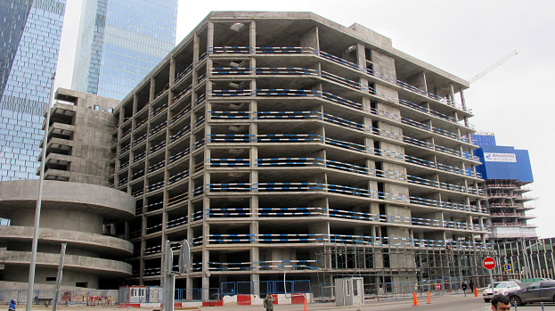 Großes Bauwerk im Bau in einer Stadt umgeben von Wolkenkratzern, Fahrzeugen, Fußgängern, Verkehrsinfrastruktur und Kränen unter einem klaren Himmel.