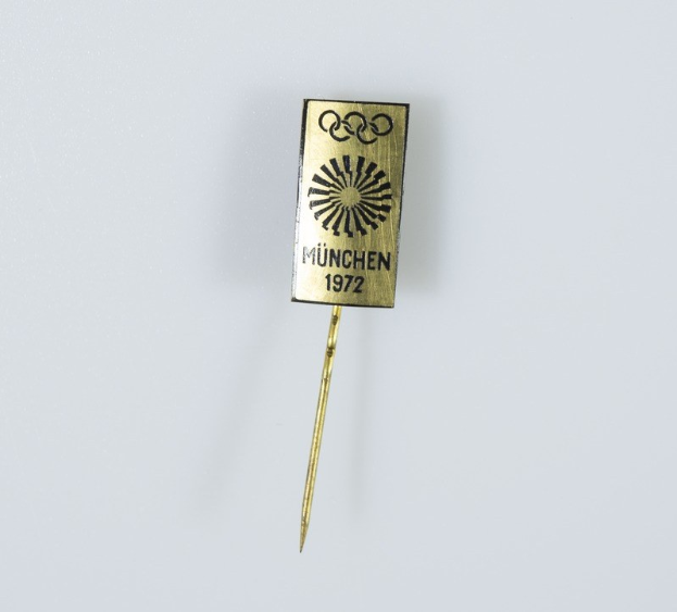 Goldene Anstecknadel mit «München 1972 Olympische Spiele» Text auf weißem Hintergrund.