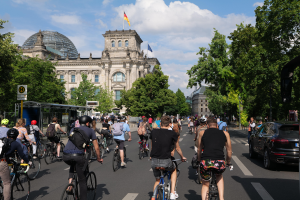 Eine Gruppe von Menschen, die Fahrräder auf einer von Bäumen gesäumten Straße in Berlin, Deutschland, fahren, mit Gebäuden, einem Bushaltestellenschild auf der rechten Seite und einer Flagge, die auf einem der Gebäude weht, unter einem bewölkten Himmel.