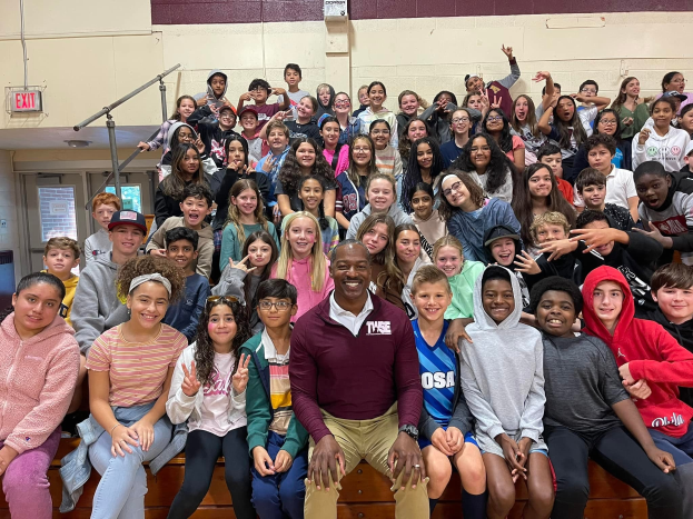 Gruppe von Schülern, die für ein Foto in einer Turnhalle posieren, einige sitzen auf der Treppe und andere stehen, eine Tür und ein Ausgangsschild auf der linken Seite sichtbar, was auf ein Sommerlager hindeutet.