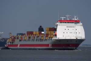 Ein großes rotes und weißes Containerschiff fährt auf dem Wasser mit einem Schlepper im Vordergrund, Containern, Bäumen und dem Himmel im Hintergrund und einem Wasserzeichen in der rechten unteren Ecke.