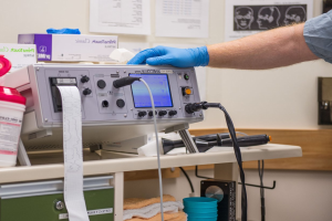 Eine Person in blauen Handschuhen arbeitet an einem medizinischen Gerät auf einem Tisch, der mit verschiedenen Gegenständen übersät ist, mit Papieren an der Wand im Hintergrund, was auf eine Krankenhausumgebung hindeutet.