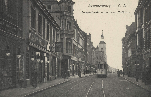 Schwarz-weiß-Foto einer Stadtstraße in Brandenburg, Deutschland mit einer Straßenbahn, Fußgängern, Gebäuden, Schildern, Straßenlaternen und einem bewölkten Himmel.