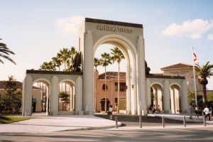 Eingang zum Universal Studios Hollywood mit einem Bogen mit Text, einer Statue, Menschen, Bäumen, Gebäuden, einem Fahnenmast mit einer Fahne.