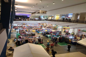 Eine feierliche Menge schlendert durch ein Einkaufszentrum mit Ständen, aufgehängten Kleidern, dekorativen Fahnen, Luftballons, sitzenden und stehenden Menschen, Geländern, Säulen und Deckenlampen, was an einen Weihnachtsmarkt im Mall of Asia erinnert.