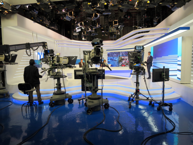 Ein TV-Studio mit mehreren Kameras auf dem Boden, einer sitzenden Person im Vordergrund und einigen stehenden Personen im Hintergrund, mit an der Decke montierten Lichtern für eine Livesendung.