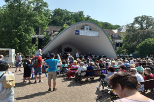 Menschenmenge auf Bänken sitzend, die einer Bühne mit stehenden Zuschauern, einer Kamera, einem Stativ, einer Fahne, einem Metallrahmen, Bäumen, Gebäuden und einem bewölkten Himmel bei einem Sommerkonzert im Park zugewandt sind.