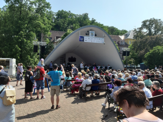 Menschenmenge auf Bänken sitzend, die einer Bühne mit stehenden Zuschauern, einer Kamera, einem Stativ, einer Fahne, einem Metallrahmen, Bäumen, Gebäuden und einem bewölkten Himmel bei einem Sommerkonzert im Park zugewandt sind.